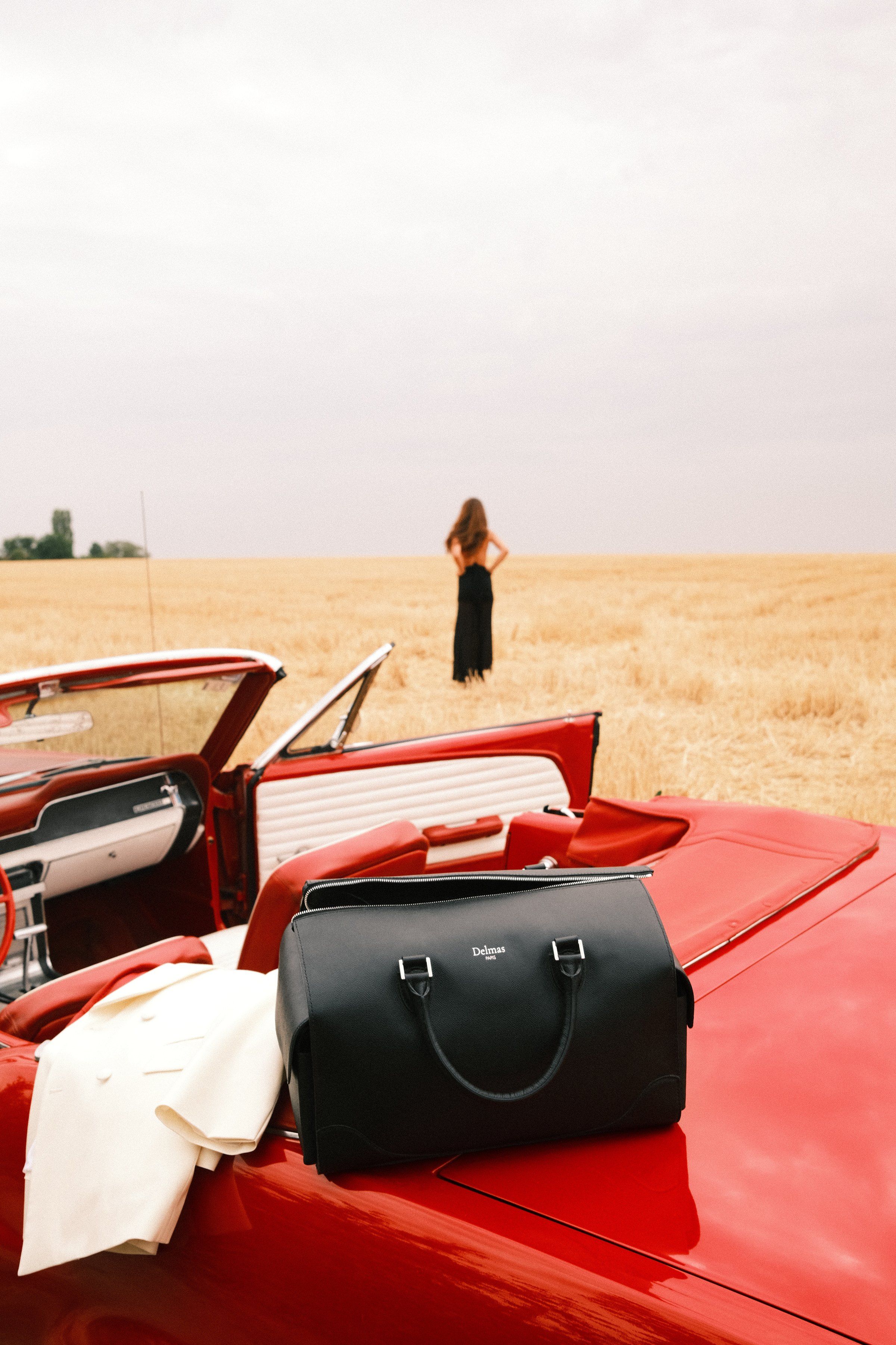 Le Baisenville noir posé sur le coffre d'une Mustang rouge, au bord d'un champ de blé. Au loin, une femme en robe noire.